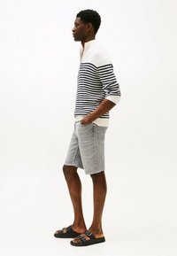 Gebreide pullover met marineblauwe en witte strepen, gecombineerd met grijze denim shorts. Zwarte sandalen met dubbele bandaccenten maken de outfit compleet.