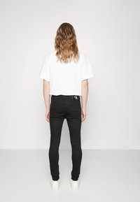 Jeans slim noirs à surface texturée, associés à un t-shirt blanc uni. Vue de dos, avec un logo sur la ceinture.