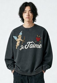Jeune personne aux cheveux noirs mi-longs portant un sweat-shirt noir orné d'un chérubin avec un arc et des flèches, un cœur rouge et le texte "Je T’aime".