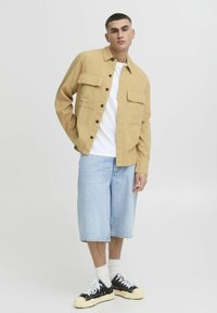 Veste beige à boutons avec deux poches sur la poitrine, portée sur un t-shirt blanc, accompagnée d'un short en jean bleu clair et de baskets noires. Design épuré.