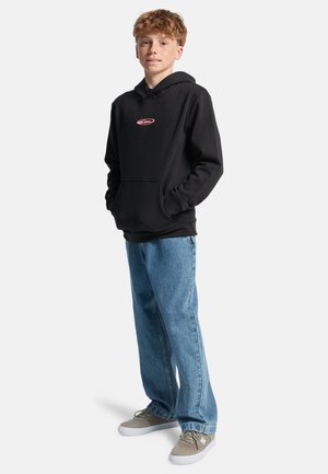 Teenager mit roten Haaren, der einen schwarzen Hoodie mit pinkem Logo, blaue Jeans und graue Sneakers trägt und mit den Händen in den Taschen steht.