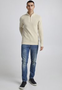 Beiger Strickpullover mit einem Viertel-Reißverschluss, gerippter Textur und figurbetontem Design. Kombiniert mit hellblauen Jeans und schwarzen Sneakers.
