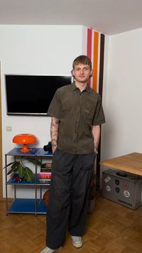 Νεαρός άνδρας με τατουάζ, που φοράει πουκάμισο ελιάς και σκουρόχρωμο παντελόνι, στέκεται σε ένα δωμάτιο με τηλεόραση, πορτοκαλί λάμπα, φυτά και μια βαλίτσα καλυμμένη με αυτοκόλλητα.