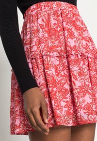 Röd blommönstrad kjol med samlad design och volangkant. Tillverkad av lättviktsmaterial med en rosa bakgrund och elastisk midja.