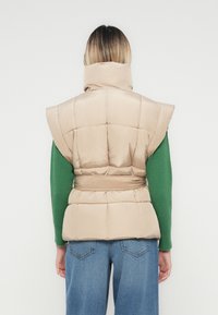 Ženska s ramenskim dolžinom las, ki nosi bež napihnjeni jopič z visokim ovratnikom, zeleni dolg rokav in modre kavbojke, prikazana s hrbta.