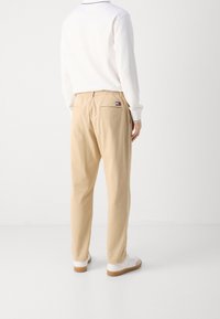 Osoba ubrana w beżowe spodnie z elastycznym pasem i małym logo na tylnej kieszeni, w połączeniu z białymi trampkami i białą bluzką z długim rękawem.