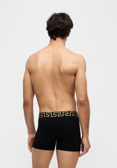Homme debout de dos, portant un boxer noir avec une ceinture ornée d’un motif clé grecque doré sur un fond clair uni.