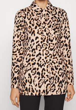 Femme portant un chemisier boutonné à manches longues avec un motif léopard beige, noir et marron et un pantalon noir.