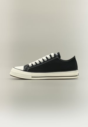 Black low-top canvas sneaker with white laces, white rubber toe cap, and white sole featuring black stripes, shown in profile on neutral background.