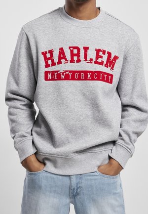 Person trägt hellgrauen Sweatshirt mit rotem Schriftzug „HARLEM“ und „NEW YORK CITY“, Hände in den Taschen, kombiniert mit hellblauen Jeans.