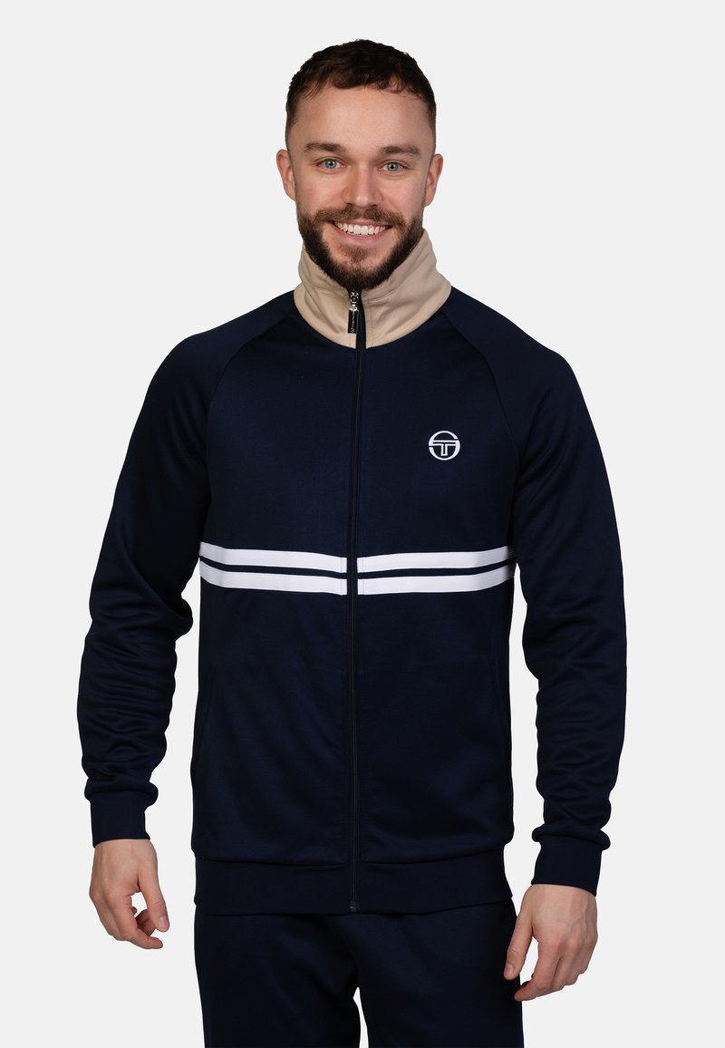 Marineblaue Zip-Jacke mit beigem Kragen, ausgestattet mit weißen Horizontalstreifen und einem runden Logo auf der Brust. Glattes, dehnbares Material.