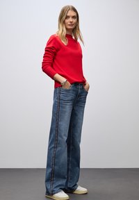 Suéter rojo, vaqueros anchos azules con tiras laterales y zapatillas blancas. El conjunto presenta un ajuste relajado y una textura suave.