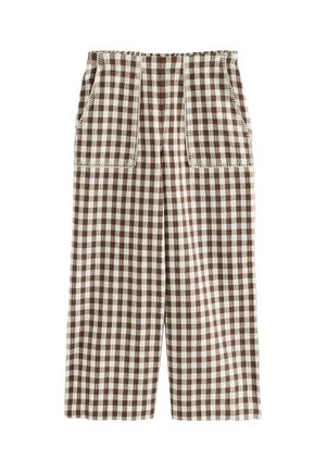 Vrouw staande op het dek van een zeilboot met een bruin-wit geruit mouwloos topje en broek met bruine sandalen onder een heldere hemel.