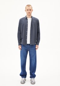 Gestreifte Herrenhemd in Marineblau mit weißen vertikalen Streifen, kombiniert mit lockeren blauen Jeans und grauen Sneakern. Weißes T-Shirt unter dem Hemd.