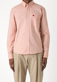 Hellrosa Button-up-Hemd mit einer Brusttasche und kleinem rotem Logo, aus einem glatten, strukturierten Stoff, kombiniert mit hellbeigen Hosen.