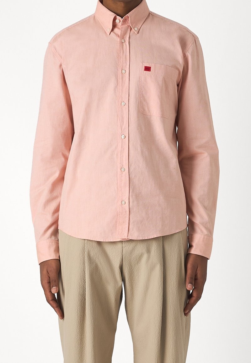 Hellrosa Button-up-Hemd mit einer Brusttasche und kleinem rotem Logo, aus einem glatten, strukturierten Stoff, kombiniert mit hellbeigen Hosen.