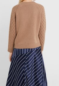 Brązowy sweter dziany z rękawami w splocie warkoczowym, ściągaczowymi mankietami i okrągłym dekoltem. Nosi się go na niebieskiej spódnicy z białymi pionowymi paskami.