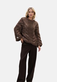 Brązowy sweter dziany ze wzorem warkocza, szerokimi mankietami i luźną sylwetką, w połączeniu z ciemnymi spodniami o szerokich nogawkach. Szczegóły z fakturowanego materiału.