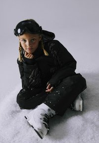 Schwarze Winterjacke mit grauen Mustern, schwarze Skibrille und weiße Skistiefel. Sitzt im Schnee mit Schneeflocken auf der Kleidung und dem Boden.