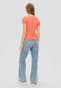 Geribbelde koraalkleurige top met korte mouwen, gecombineerd met lichtblauwe wijde jeans. De jeans hebben achterzakken en een relaxte pasvorm, met een eenvoudige witte schoen.