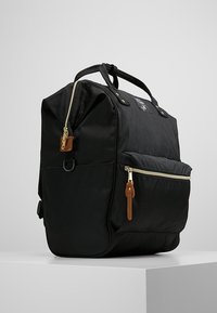 Sac à dos noir de forme rectangulaire, fabriqué en tissu durable. Doté de fermetures éclair dorées, de détails en cuir marron, et de deux poignées supérieures.
