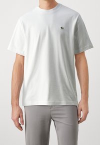 Bílá bavlněná tričko s krátkými rukávy a designem s kulatým výstřihem, s malým zeleným logem Lacoste na levé hrudi. Hladká textura.