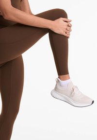 Barna leggings sima textúrával, világos bézs sportcipővel, kötött felsőrésszel és fehér talppal, fehér háttér előtt.