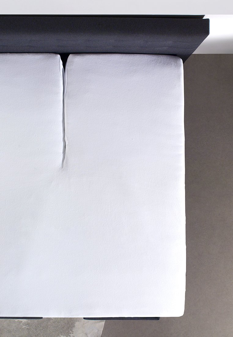 Två vita kuddar med en slät textur ligger intill ett svart horisontellt sänggavel på en grå yta. Enkla, moderna designelement är närvarande.