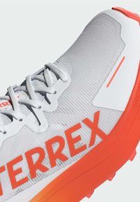 Lette grå sneakers med en mesh-overdel, klare orange accenter og iøjnefaldende "TERREX" branding på siden. Tekstureret design med snørebånd.