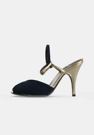Sandalia de tacón alto peep-toe negra con tacón de aguja dorado y correa en el tobillo sobre un fondo claro y liso.