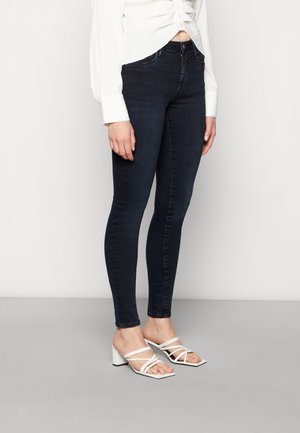 Frau trägt dunkle Skinny-Jeans, weiße langärmelige Bluse und weiße Sandalen mit mehreren Riemen, steht auf grauem Boden.