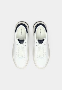 Tênis de couro branco com detalhes em azul-marinho, bico redondo, atacadores planos e sola almofadada. Logótipo "GUESS" em destaque na língua e na lateral.