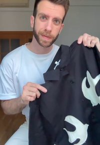 Camisa preta com impressão grande de uma pomba branca, diseño de botões e colarinho, segurada por uma pessoa vestindo uma t-shirt branca. Textura suave, tecido de peso médio.