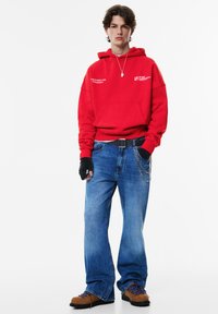 Punainen huppari, jossa etutasku ja teksti, yhdistettynä sinisiin leveälahkeisiin farkkuihin, mustiin hansikkaisiin ja ruskeisiin kenkiin, joissa siniset nauhat. Hopeinen ketjuyksityiskohta.