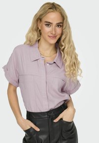 Camisa corta de manga corta de color púrpura claro con diseño de botones, que cuenta con dos bolsillos frontales y un ajuste relajado, combinada con pantalones de cuero negros.