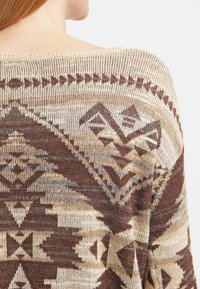 Stickad tröja i beige med geometriska mönster i brunt och grått. Mjuk textur, bred halsringning och design med öppen rygg.