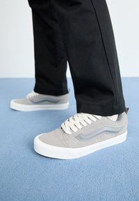 Szare zamszowe sneakersy z białymi gumowymi podeszwami, z szarymi paskami po bokach i białymi sznurowadłami, zestawione z czarnymi spodniami na niebieskiej powierzchni.