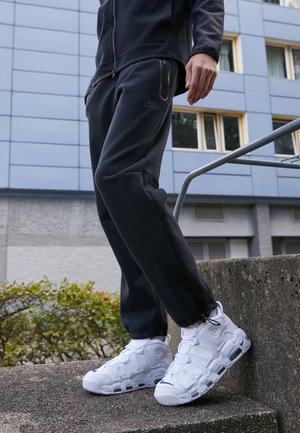 Osoba ubrana w czarne spodnie dresowe i białe buty sportowe Nike stoi na zewnętrznych betonowych schodach w pobliżu poręczy i elewacji budynku.
