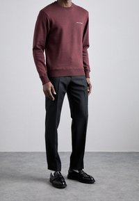 Uomo che indossa una felpa bordeaux con piccolo testo bianco, pantaloni neri su misura con tasche, calzini bianchi e mocassini neri su un pavimento grigio.