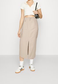 Jupe cargo beige avec poches latérales et fente avant, associée à un haut court blanc et des baskets blanches à rayures noires.