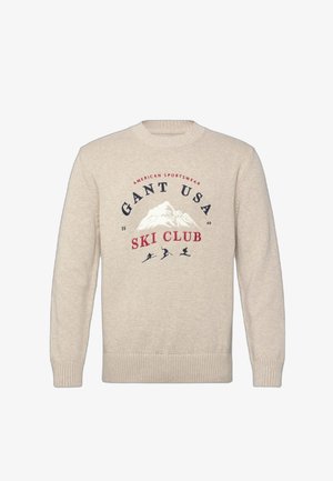 Maglione beige a maglia con grafica di montagna e testo "Gant USA Ski Club," con tre silhouette di sciatori sotto il disegno.