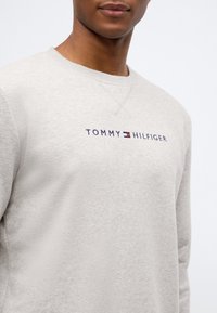 Sweatshirt gri deschis, din material moale, cu guler rotund și logo-ul "TOMMY HILFIGER" în nuanțe de bleumarin și roșu pe față.