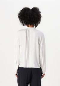 Blusa bianca a maniche lunghe con scollo posteriore arricciato e tessuto morbido e liscio. Abbinata a pantaloni scuri, vista da dietro.