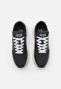 Schwarze Turnschuhe mit weißen Schnürsenkeln, die eine Kombination aus Leder- und Textilmaterialien aufweisen. Die Sohle ist weiß mit einem gesprenkelten Muster. Markenlogo vorhanden.