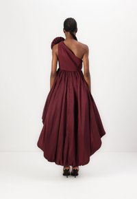 Burgundy-henkinen mekko, jossa on rakenteellinen frilla olkapäällä, tiukka vartalonmyötäinen yläosa ja runsas hame, joka laskeutuu nilkkoihin saakka.