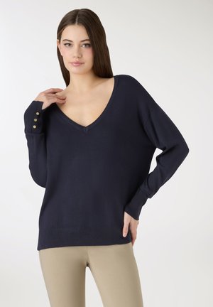 Giovane donna con capelli castani lisci che indossa un maglione blu navy a scollo a V con bottoni dorati su un polsino e pantaloni beige.