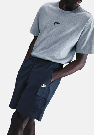 Mężczyzna w szarej koszulce Nike i czarnych szortach cargo Nike, ręce w kieszeniach, na tle jednolitej białej ściany.