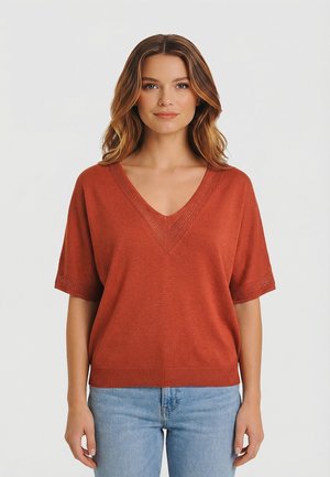 Mujer con cabello suelto y ondulado, vestida con un suéter de manga corta y cuello en V de color óxido y jeans azul claro, de pie frente a un fondo liso y claro.