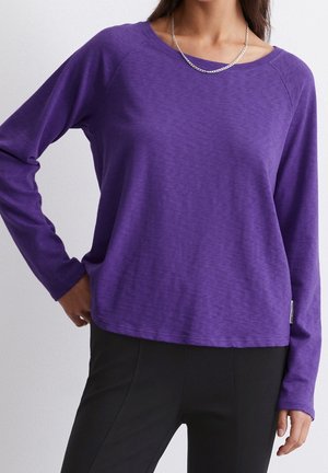 Frau trägt ein lila langärmliges Hemd und schwarze Hose, steht mit einer Hand in der Hüfte vor einem einfarbigen hellen Hintergrund.