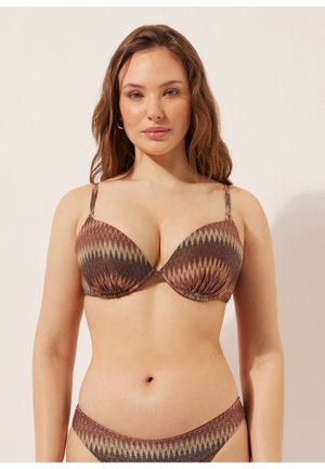 Ung kvinde iført en brun bikini med zigzag-mønster, stående mod en ensfarvet lys baggrund, vendt fremad med afslappet udtryk.